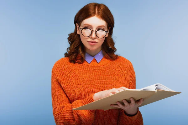 Estudiante pelirroja en gafas y suéter naranja sosteniendo libro y mirando hacia otro lado aislado en azul - foto de stock