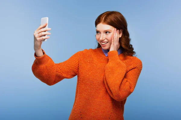 Giovane donna sorridente in maglione arancione prendendo selfie isolato su blu — Foto stock
