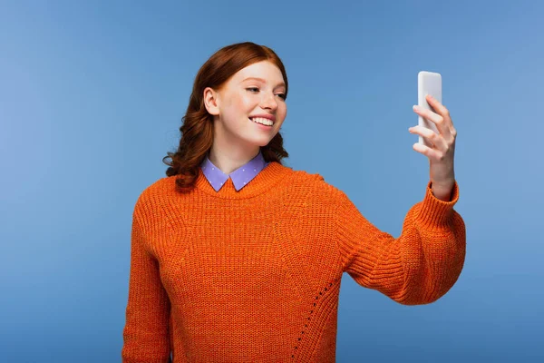 Smiling young woman in orange sweater taking selfie isolated on blue — Stock Photo