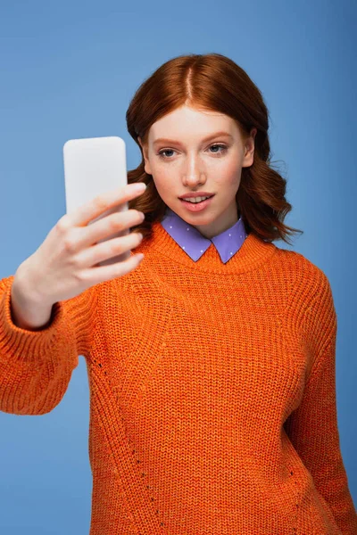 Рыжая женщина в оранжевом свитере делает селфи, изолированное на голубом — стоковое фото