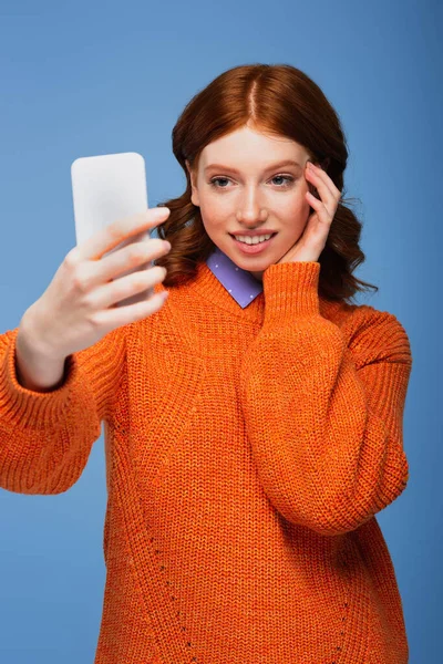 Happy redhead woman in orange sweater taking selfie on smartphone isolated on blue — Stock Photo