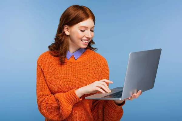Felice studentessa rossa in maglione arancione utilizzando computer portatile isolato su blu — Foto stock