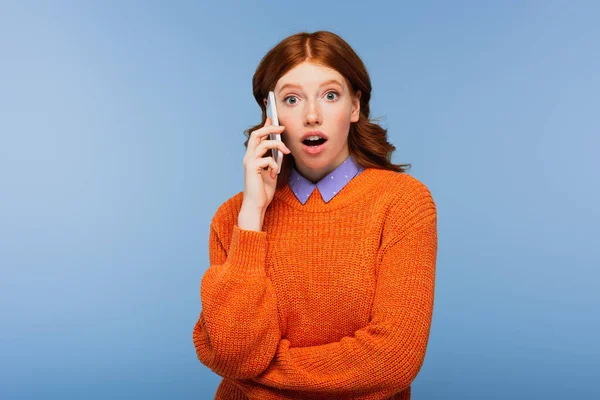 Mujer pelirroja sorprendida en suéter naranja hablando en teléfono inteligente aislado en azul - foto de stock