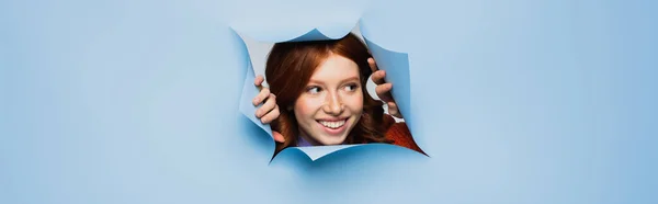 Cheerful redhead young woman looking away on blue ripped background, banner — Stock Photo