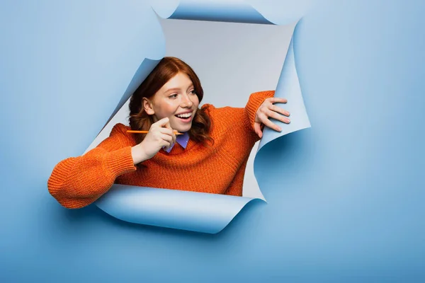 Happy young woman in orange sweater holding pencil and looking away on blue ripped background — Stock Photo