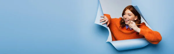 Jovem feliz em suéter laranja segurando lápis e olhando para longe no fundo rasgado azul, banner — Fotografia de Stock