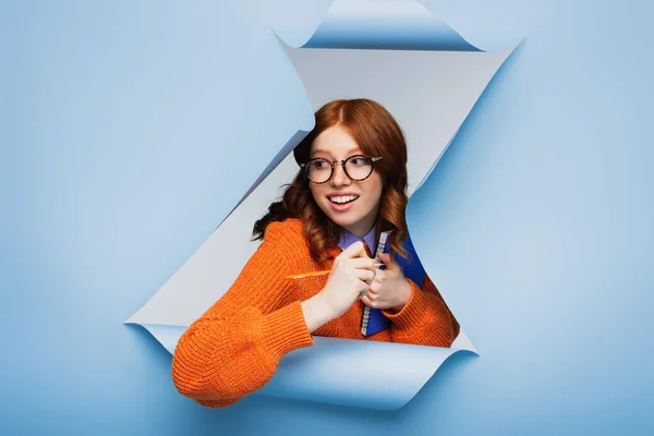 Rousse jeune femme en pull orange et lunettes tenant carnet et crayon sur fond bleu déchiré — Photo de stock