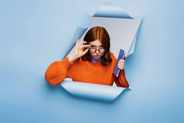 Rousse jeune étudiant en pull orange et lunettes tenant carnet et crayon sur fond bleu déchiré — Photo de stock