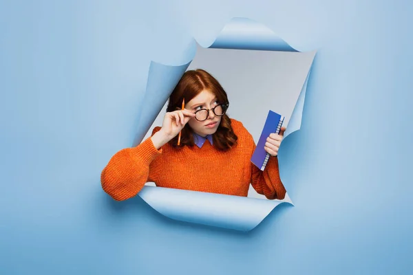 Pelirroja joven estudiante en suéter naranja y gafas sosteniendo bloc de notas y lápiz sobre fondo azul rasgado - foto de stock