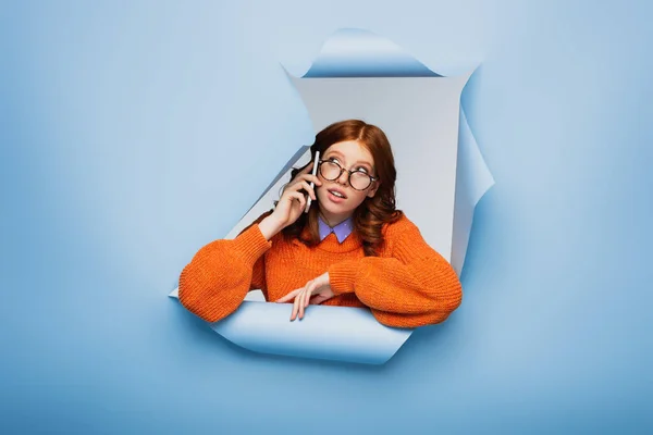 Redhead young woman in orange sweater adjusting glasses and talking on cellphone on blue ripped background — Stock Photo