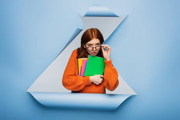 Ruiva jovem estudante em suéter laranja ajustando óculos e segurando cadernos em azul rasgado fundo de papel — Fotografia de Stock