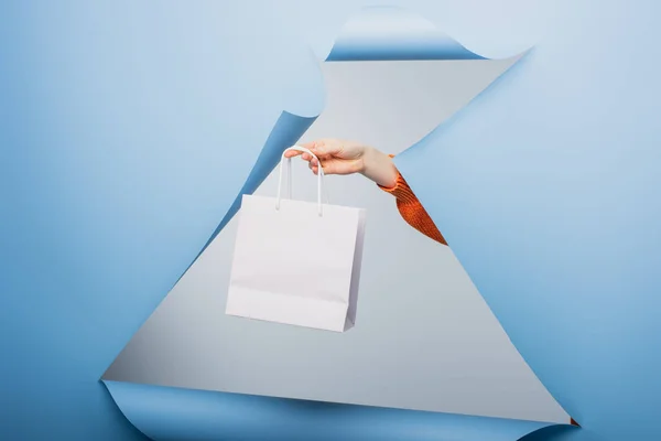 Vue recadrée d'une femme tenant un sac à provisions blanc sur fond de papier déchiré bleu — Photo de stock