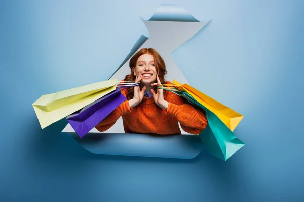 Happy redhead woman holding colorful shopping bags on blue ripped paper background — Stock Photo