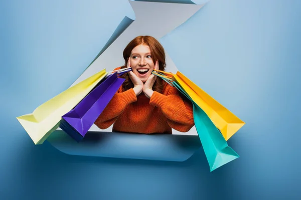 Excité rousse femme tenant des sacs colorés shopping sur fond de papier déchiré bleu — Photo de stock