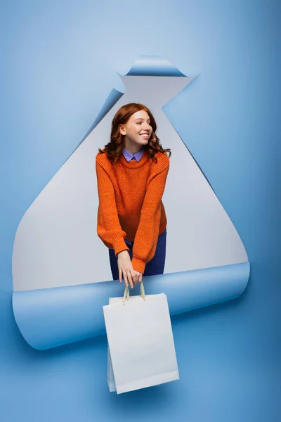 Улыбающаяся рыжая женщина держит сумку с покупками на голубом разорванном бумажном фоне — стоковое фото