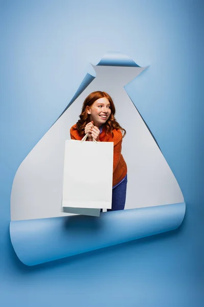 Smiling young woman holding shopping bag on blue ripped paper background — Stock Photo