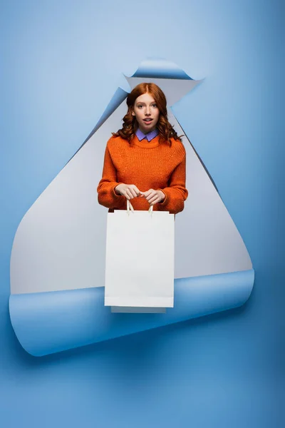 Surpresa jovem segurando saco de compras no fundo de papel rasgado azul — Fotografia de Stock