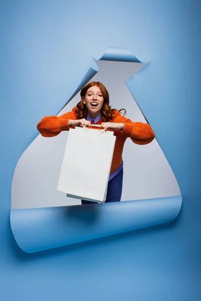 Mujer joven asombrada sosteniendo bolsa de compras sobre fondo de papel rasgado azul - foto de stock