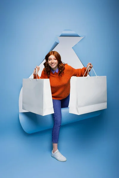 Rossa positiva giovane donna che tiene borse della spesa su sfondo di carta strappata blu — Foto stock