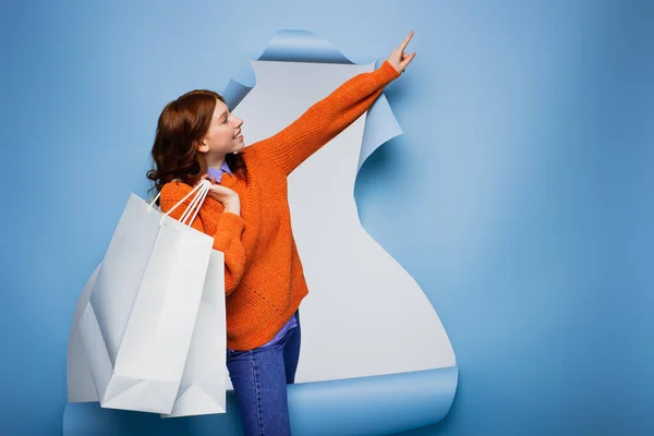 Jeune femme heureuse tenant des sacs à provisions et pointant du doigt sur fond de papier bleu déchiré — Photo de stock
