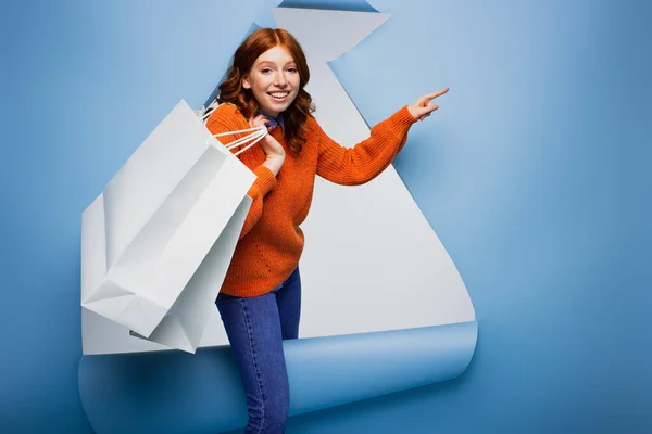 Joven mujer sonriente sosteniendo bolsas de compras y señalando con el dedo sobre fondo de papel rasgado azul - foto de stock
