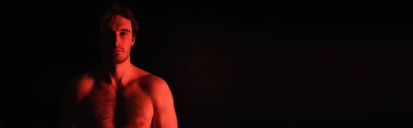 Illuminazione rossa su sexy uomo senza maglietta guardando la fotocamera isolata su nero, banner — Foto stock
