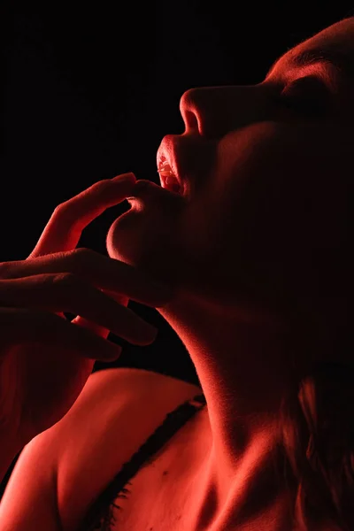 Luz roja en la mujer seductora tocando los labios aislados en negro - foto de stock