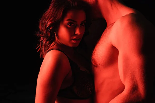 Червоне освітлення на сексуальній жінці в нижній білизні біля м'язового чоловіка ізольовано на чорному — стокове фото