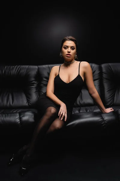 Longitud completa de la mujer seductora en vestido de deslizamiento sentado en sofá de cuero en negro — Stock Photo