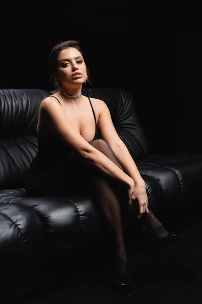 Comprimento total da mulher sedutora em vestido de deslizamento e meias sentadas no sofá em preto — Fotografia de Stock