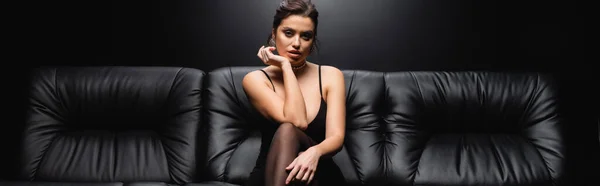 Сексуальная женщина в скользящем платье и колготки сидя на диване на черном фоне, баннер — стоковое фото