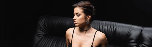 Sexy donna in collana e orecchini seduto sul divano isolato su nero, banner — Foto stock