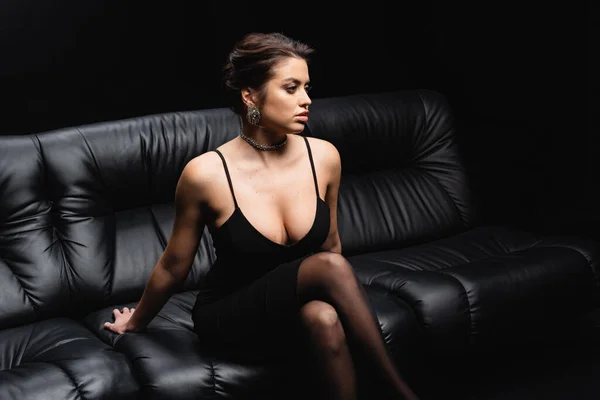 Sexy woman in slip dress and tights sitting on leather sofa isolated on black — Stock Photo