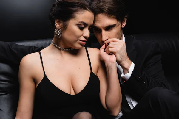 Passionate man in suit kissing hand of seductive woman in slip dress on black — Stock Photo