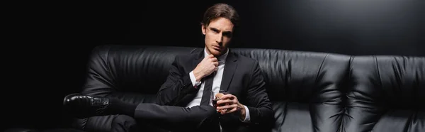 Elegante uomo in giacca e cravatta seduto su divano in pelle con bicchiere di whisky su sfondo nero, banner — Foto stock