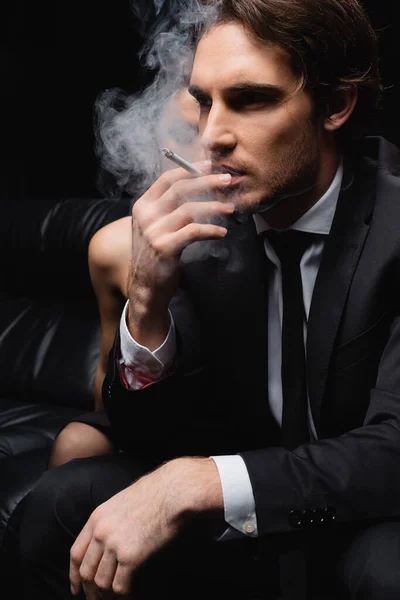 Серьезный мужчина в костюме курит сигарету рядом с женщиной на размытом черном фоне — стоковое фото
