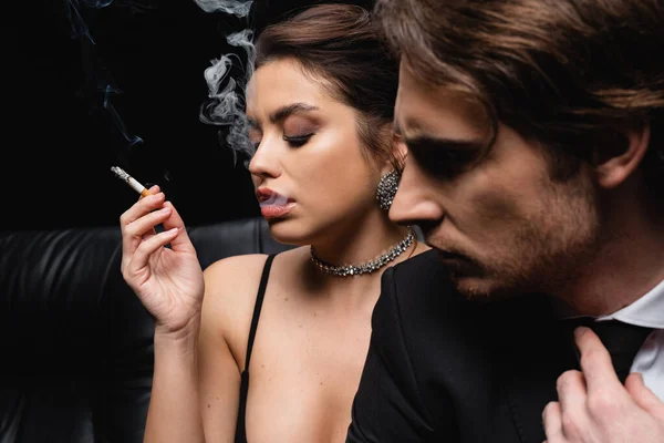 Жінка в сережках і намистах курить сигарету біля чоловіка на розмитому фоні — стокове фото