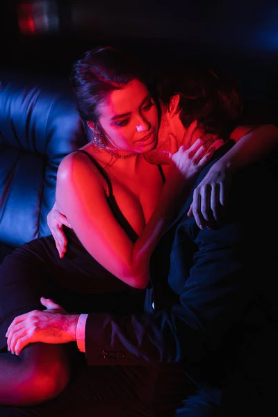 Red and blue lighting on sexy couple hugging and kissing on black leather sofa — Stock Photo