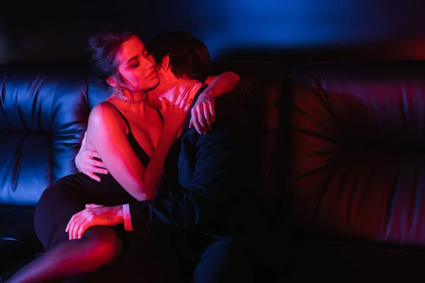 Iluminación roja y azul en el hombre besar a la mujer sexy en el sofá de cuero negro - foto de stock