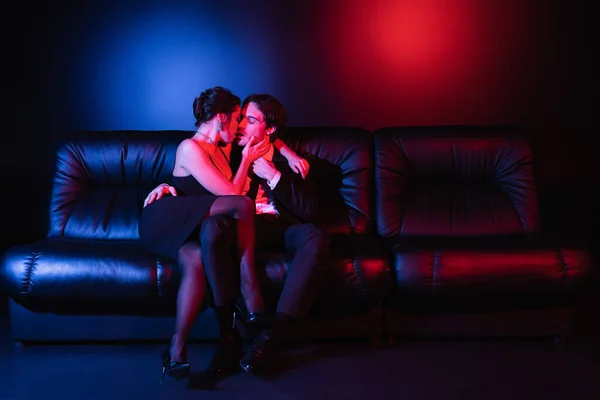 Full length of red and blue lighting on sexy man and woman kissing on black leather sofa — Stock Photo