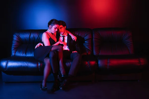 Pleine longueur d'éclairage rouge et bleu sur couple sexy étreignant sur canapé en cuir noir — Photo de stock