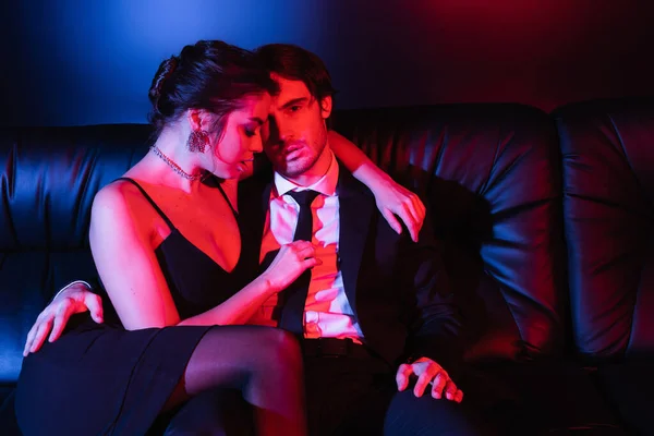 Blue and red lighting on sexy couple embracing on black leather sofa — Stock Photo