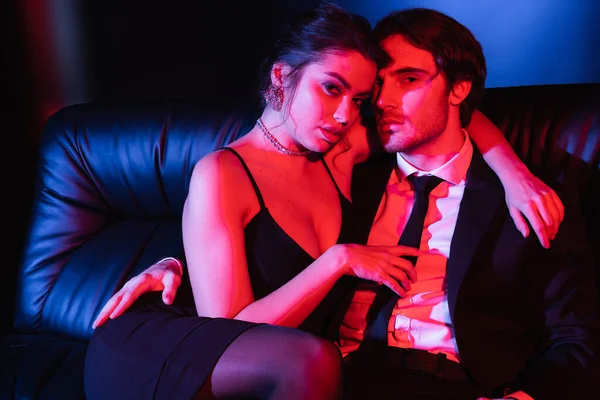 Iluminación azul y rojo en pareja sexy abrazándose en sofá de cuero negro - foto de stock