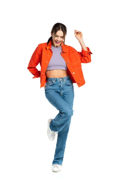 Piena lunghezza di positivo giovane donna in crop top, jeans e camicia arancione posa isolata su bianco — Foto stock