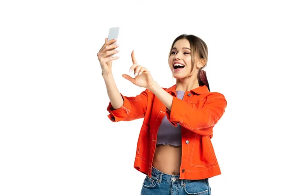 Stupito giovane donna in crop top e camicia arancione prendendo selfie su smartphone e gesticolando isolato su bianco — Foto stock