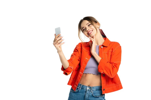 Mujer joven positiva en la parte superior de la cosecha y camisa naranja tomar selfie en el teléfono inteligente aislado en blanco - foto de stock