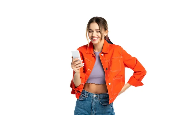 Mujer joven feliz en la parte superior de la cosecha y camisa naranja usando el teléfono inteligente, mientras que de pie con la mano en la cadera aislado en blanco - foto de stock