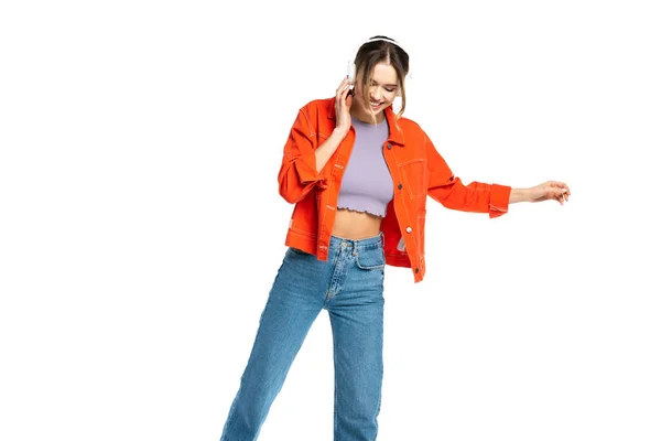 Glückliche Frau in Jeans, bauchfreiem Top und orangefarbener Jacke, die Musik in drahtlosen Kopfhörern isoliert auf weiß hört — Stockfoto