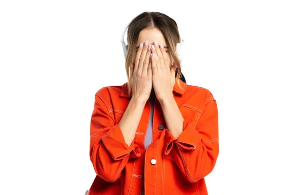 Mulher de camisa laranja cobrindo rosto enquanto as mãos enquanto ouve música em fones de ouvido sem fio isolados no branco — Fotografia de Stock