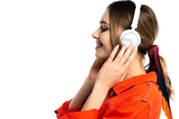 Vista laterale di donna gioiosa in camicia arancione ascoltare musica in cuffie wireless isolato su bianco — Foto stock